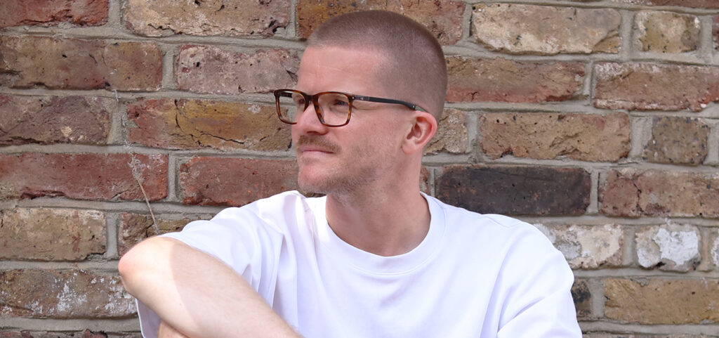 A portrait shot of a young man with a buzz cut, sitting with one arm resting casually on his knee. He is wearing a plain white tee and a pair of square sports glasses which wrap behind his ears. The background is a brick wall of mixed beige and red. 