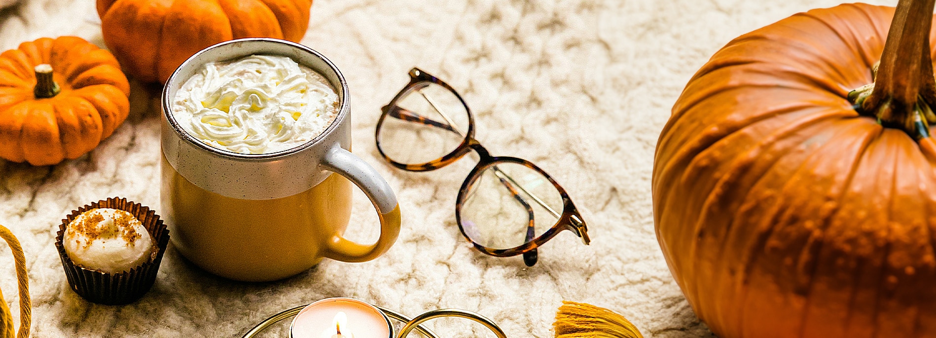 A pair of round tortoiseshell glasses rests folded on a white knitted blanket, surrounded by pumpkins, a cupcake and creamy hot drink in a ceramic mug.