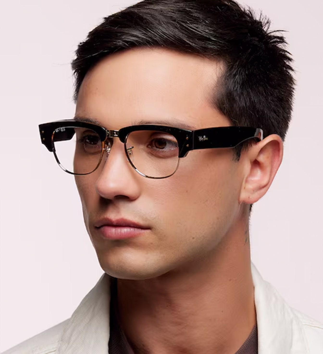 A young man with short dark hair is wearing a chunky black Ray-Ban Clubmaster frame. He appears to be wearing a white jacket over a grey top. 