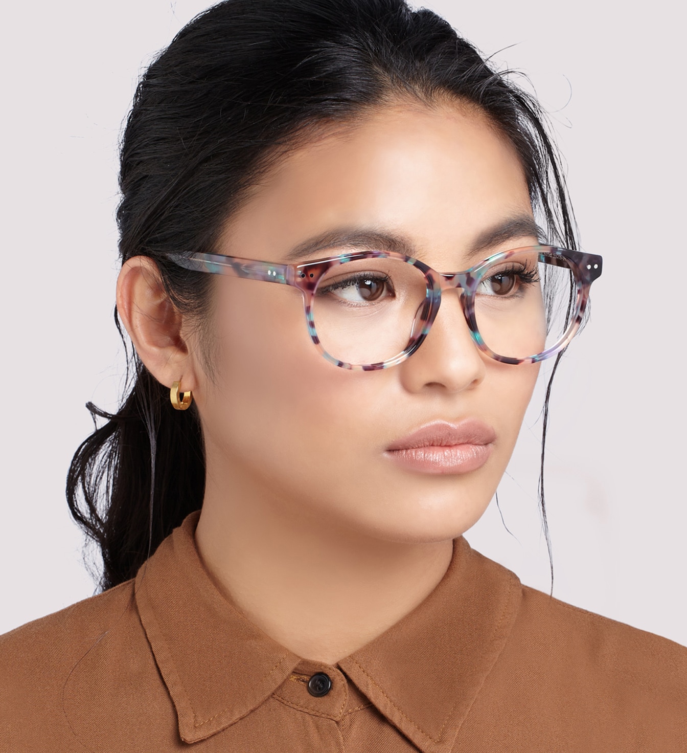 A young woman with dark hair tied in a loose ponytail wears a brightly-coloured pair of round acetate glasses. She has small gold-hooped earrings and is wearing a brown shirt. 