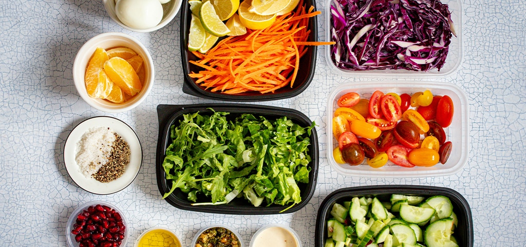 A selection of healthy foods laid out in bowls and containers, including boiled eggs, chopped carrots and limes, shredded red cabbage, chopped tomatoes, chopped cucumber, shredded lettuce, tangerine pieces, pomegranate seeds and several spices and sauces. 