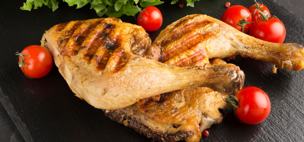 Two grilled chicken legs placed on a black slate with lettuce and cherry tomatoes garnishing the outside. 