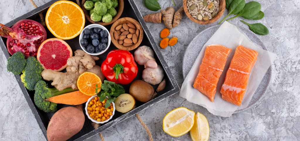 A selection of healthy foods on a grey marble surface, including two salmon fillets on grey paper and a grey plate, alongside a dark-grey wooden tray with rope handles filled with oranges, blueberries, almonds, Brussels sprouts, and other produce.