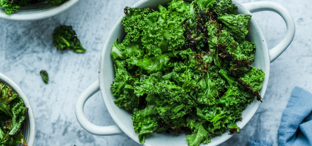 A white metal bowl with handles holding a fresh bunch of kale, placed on a white marble surface. Two additional ceramic bowls filled with leafy greens are partially visible nearby.