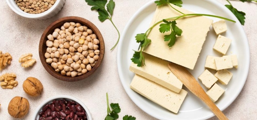 On a white kitchen surface, a brown bowl holds chickpeas beside a white plate containing a chunk of tofu, tofu slices, and a wooden spoon. A white ceramic bowl of lentils is partially visible at the top of the frame, and a separate white dish of kidney beans appears at the bottom.