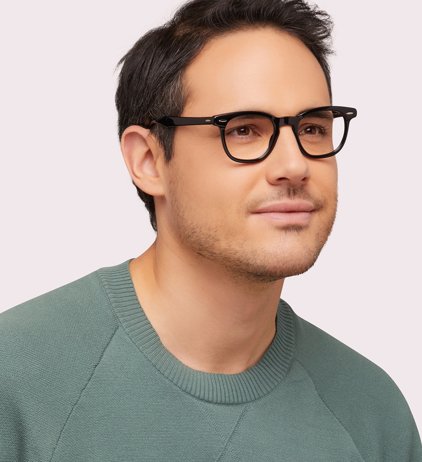 A close-up of a man with short dark hair. He is wearing a pair of black Ray-Ban glasses and a green sweater. The man is positioned at an angle. 