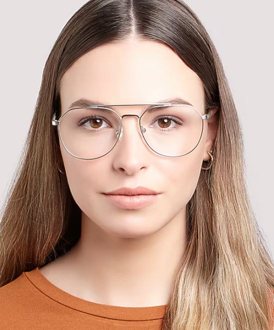 A young woman facing the camera wears gold pilot glasses and an orange top. Her long blonde hair rests on her shoulders, and she has a neutral expression against a pale background.