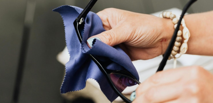 A close-up of two hands cleaning a pair of black glasses with a blue cloth. The person has blue nail varnish and is wearing two bracelets on their right hand.