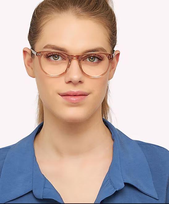 A young woman facing the camera wears round, translucent brown acetate glasses and a blue collared shirt. Her long blonde hair is tied back, and she has a neutral expression.