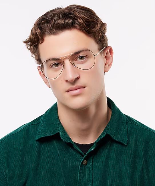 A young man facing the camera wears gold Aviator glasses and a dark green shirt. He has short wavy brown hair, and a neutral expression against a pale background.