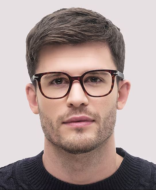A man facing the camera wears square Havana acetate glasses and a dark knitted sweater. He has short brown hair, light stubble, and a neutral expression against a pale background.