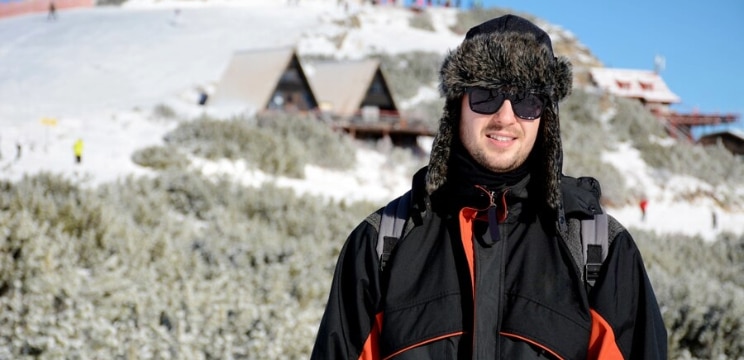 A man in a snowy alpine outdoor environment. Two cabins and snowy hills are just visible in the background. He is wearing a dark fluffy aviator hat, black sunglasses and a big black coat with red stripes. The man is smiling brightly.