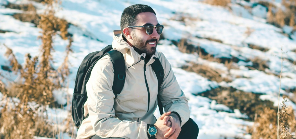 A man with short dark hair sitting in a snowy outdoor environment. He is wearing Ray-Ban Clubmaster sunglasses, a cream-coloured rain jacket, a large watch and has a black pack on his back. He is smiling brightly with his hands held together. He's looking off into the distance.