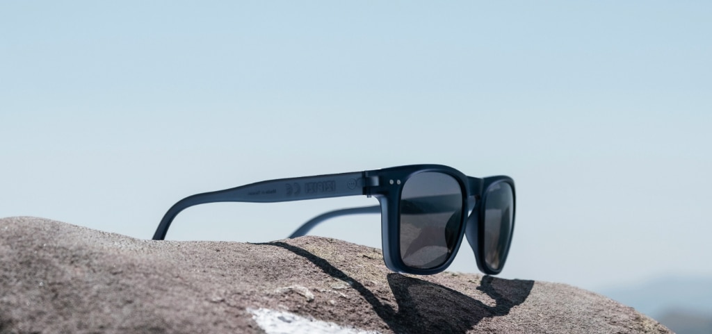 A pair of dark acetate sunglasses perched open on a grey rock with the blue sky overhead.