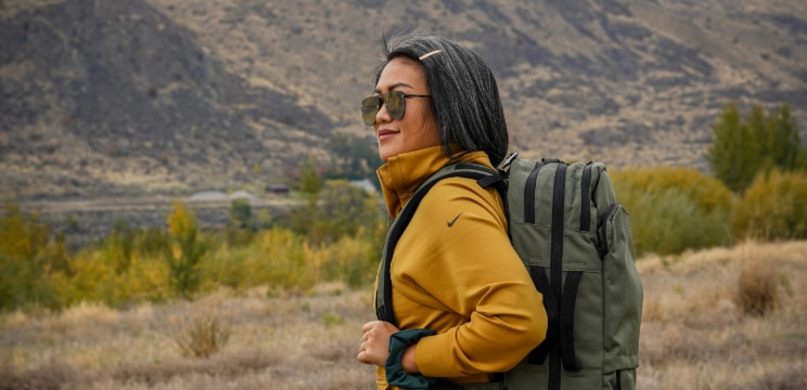 A lady looking to the left in a bright mountainous outdoor environment. She has a dark bob of hair, is wearing large sunglasses and a yellow sports jacket. The lady has a khaki backpack on her shoulders. One hand is gripping the strap of the bag.