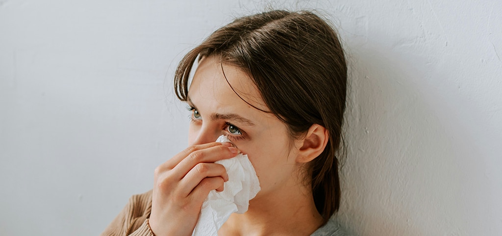 A close up of a young lady with her hair tied back. She is rubbing her eyes with a tissue and leaning against a white wall.