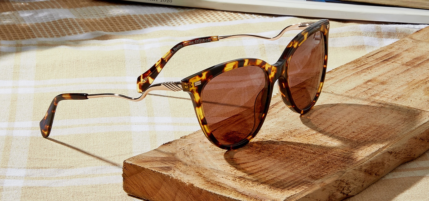 A large pair of cat-eye sunglasses in a bright yellow Havana with curvy metal temples. The sunglasses are rested on a wooden slab below which is a yellow and white picnic blanket.