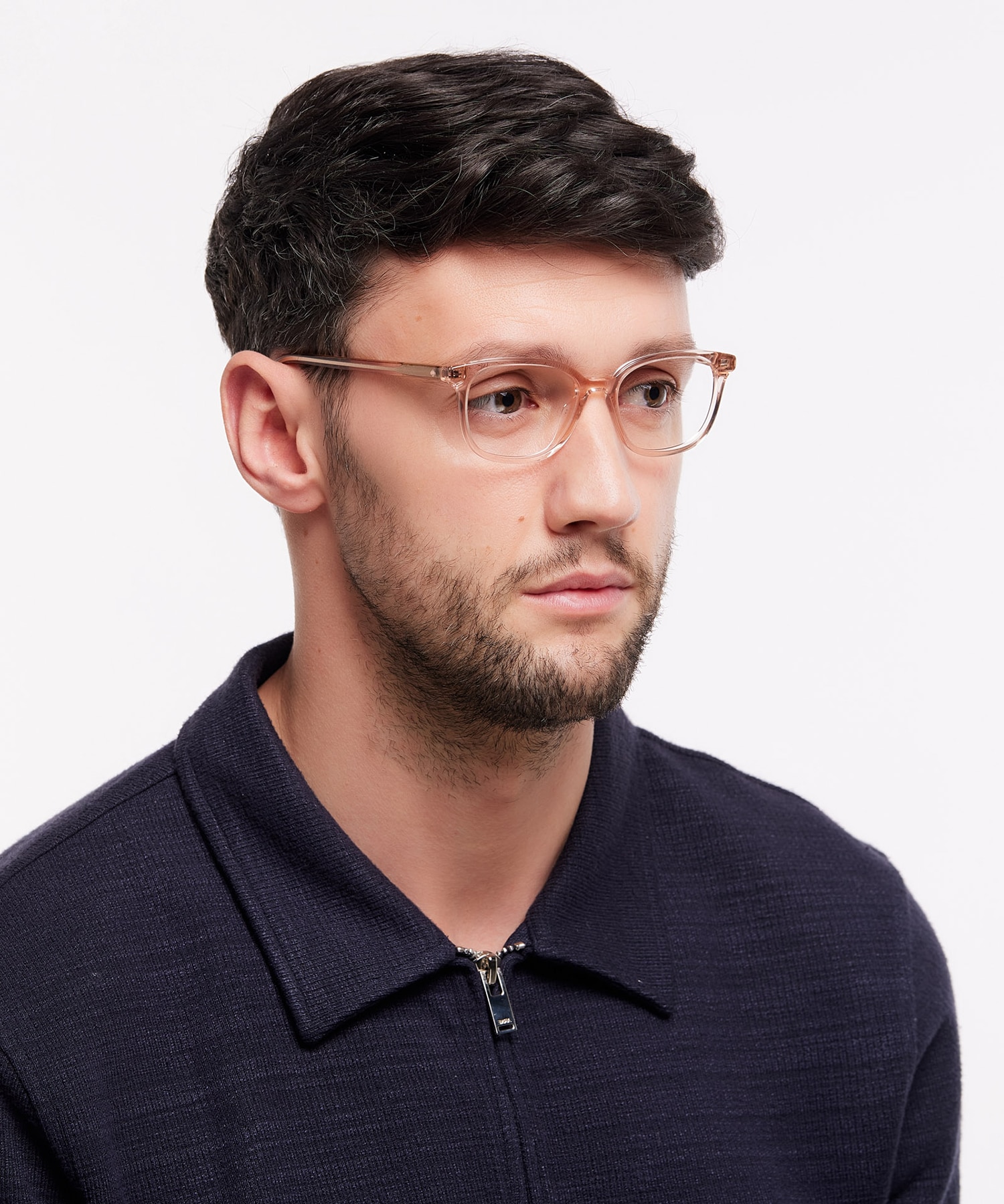 A close up of a young man with short brown hair and stubble. He is wearing a pair of clear pink acetate glasses and a navy polo shirt.