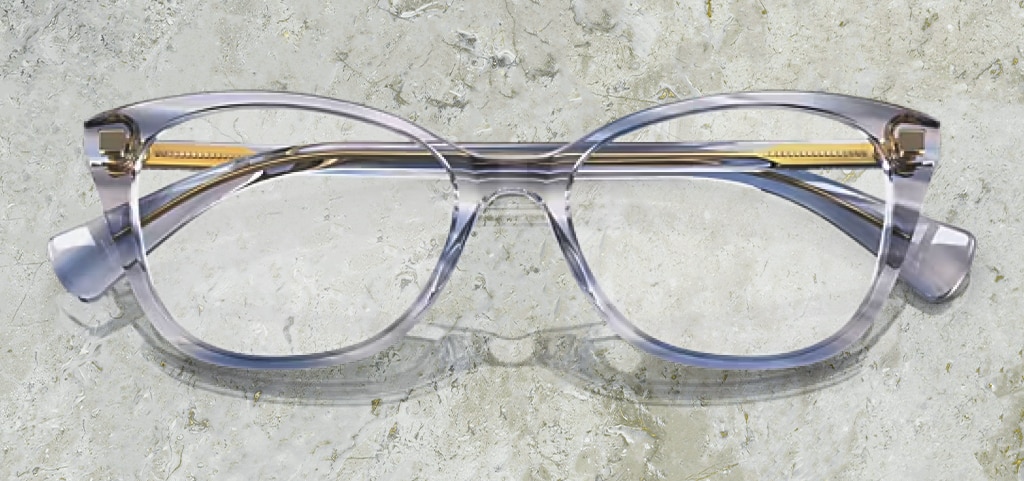 A pair of of acetate pillow-shaped Ralph by Ralph Lauren glasses. They have a transparent blue aesthetic with golden inner temples. The glasses are folded up on a marble background.