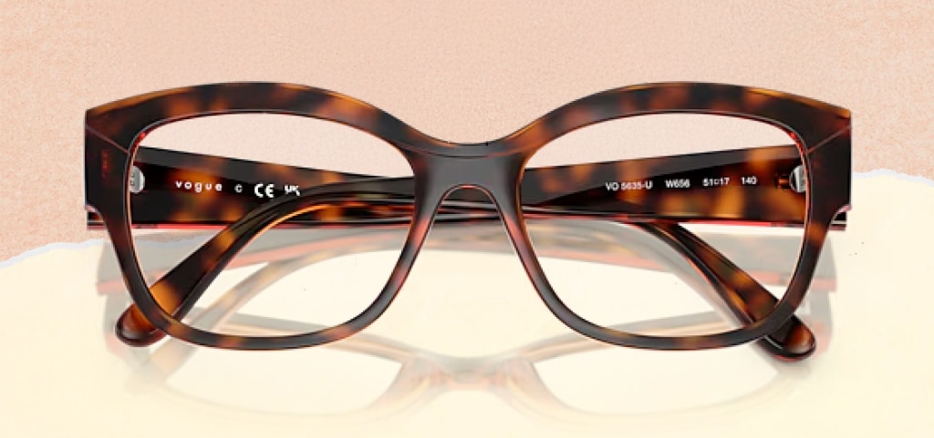 A pair of chunky pillow-shaped Vogue glasses folded up on a paper-torn peach and cream background. The glasses are a dark Havana colour.