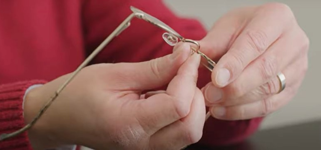 A close up of two hands adjusting the nose pads of a pair of gold metal glasses. The person is wearing a red jumper.