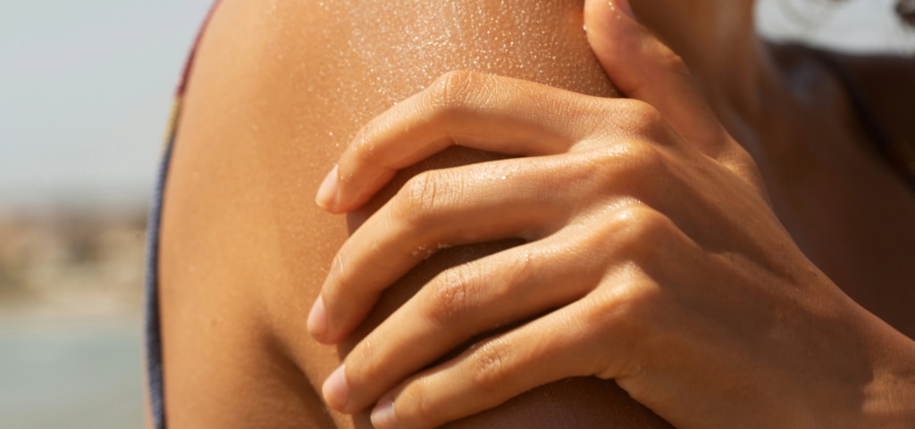 A close-up captures a woman’s shoulder as she applies lotion to her skin. She’s wearing a swimsuit, with the blurred backdrop suggesting a coastal setting.