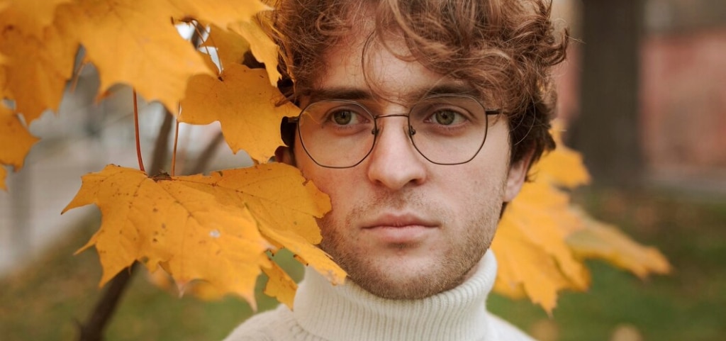A close up of a young man with curly auburn hair and hazel eyes. Orange autumnal leaves surround him. He is wearing black metal geometric glasses and a cream polo neck.