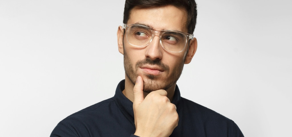 A close up of a young man with short dark hair and stubble. He is wearing transparent square glasses and a navy shirt. The young man is resting one hand on his chin and looking up to one corner as if thinking about something.
