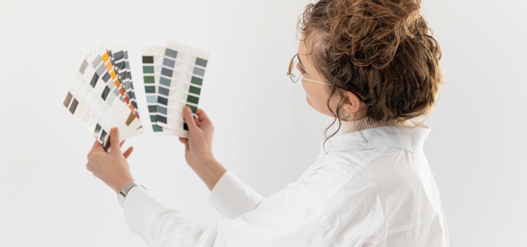 A young lady with light brown hair tied in a messy bun. She is wearing a white shirt over a cream polo neck jumper and silver metal glasses. She is facing away, looking at colour swatches which she is holding up in front of her with both hands. She appears to be comparing the colour swatches.