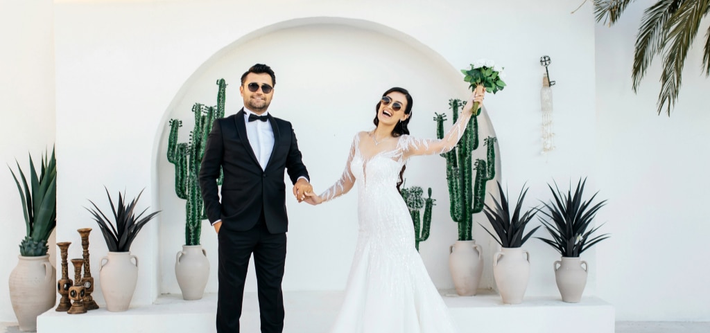 A pair of newlyweds standing in a bright white building. There are several tropical plants behind them including palm trees and cactuses. The man has short dark hair and stubble. He is wearing a black tux and black sunglasses. The lady has dark hair partially tied back. She is wearing sunglasses and a fishtail wedding dress with lace sleeves. She is holding her bouquet up in the air and smiling brightly.