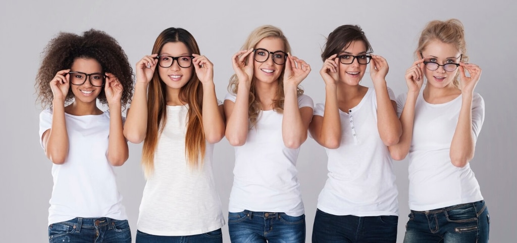 A photo of five young women with different skin tones standing in front of an off-white background. One has frizzy dark brown, medium-length hair, one has long, straight auburn hair, one has blonde curly hair, one has black hair tied back and the final lady has blonde hair tied back. They are all wearing black glasses, white t-shirts and blue jeans. They are holding their glasses frames with both hands and laughing or smiling.