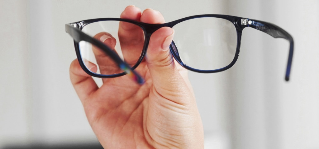 A close up of a hand holding a pair of dark blue acetate glasses in the air. The glasses arms are pointing towards the camera, allowing us to look through the lenses. The background is blurry white.