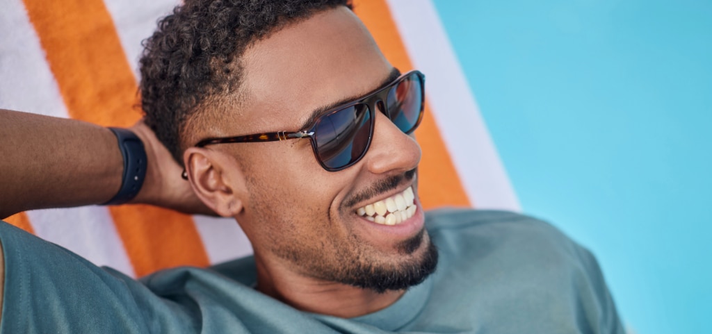 A young man lounges on a bright orange-and-white striped deck chair, one hand resting behind his head as he beams with a bright smile. His short, dark curly undercut and neat anchor-shaped beard add a stylish edge, while a light blue t-shirt and Persol Havana acetate sunglasses complete his laid-back summer look.