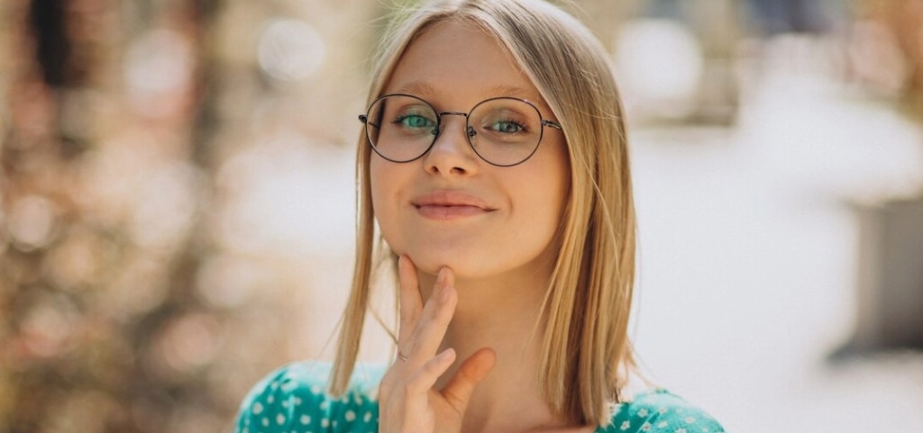 A close up of a young lady with a medium-length, blonde bob and blue eyes. She is wearing a pair of round, black metal glasses and a green floral dress. The lady is resting her right hand on her chin and smiling. The background is blurred but appears to be a bright outdoor setting.