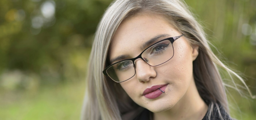 A close up of a young lady with ashy blonde long hair and grey-coloured eyes. She is wearing a pair of rectangular, black metal glasses and a black jacket. The lady has a lip piercing and is tilting her head to one side. The background is blurred but appears to be a bright outdoor setting.