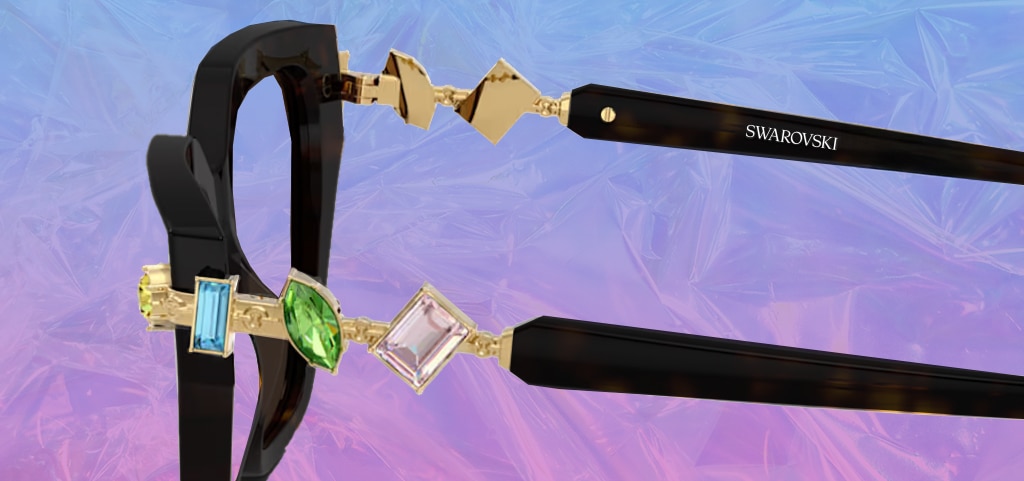 A close up of the side of a pair of black Swarovski glasses. The hinges are embellished with three gems; a blue, green and purple one. The glasses are on a shiny, reflective purple background. 