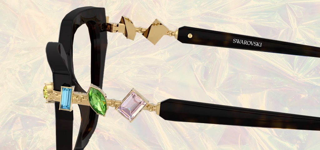 A close up of the side of a pair of black Swarovski glasses. The hinges are embellished with three gems; a blue, green and purple one. The glasses are on a shiny, reflective beige background.