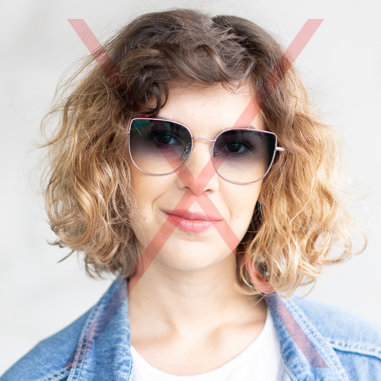 A cropped headshot of a young woman with frizzy brown hair wearing a pair of metal cat-eye sunglasses, a white t-shirt and denim top. There is a red cross through the image to indicate that it's not an appropriate passport photo.