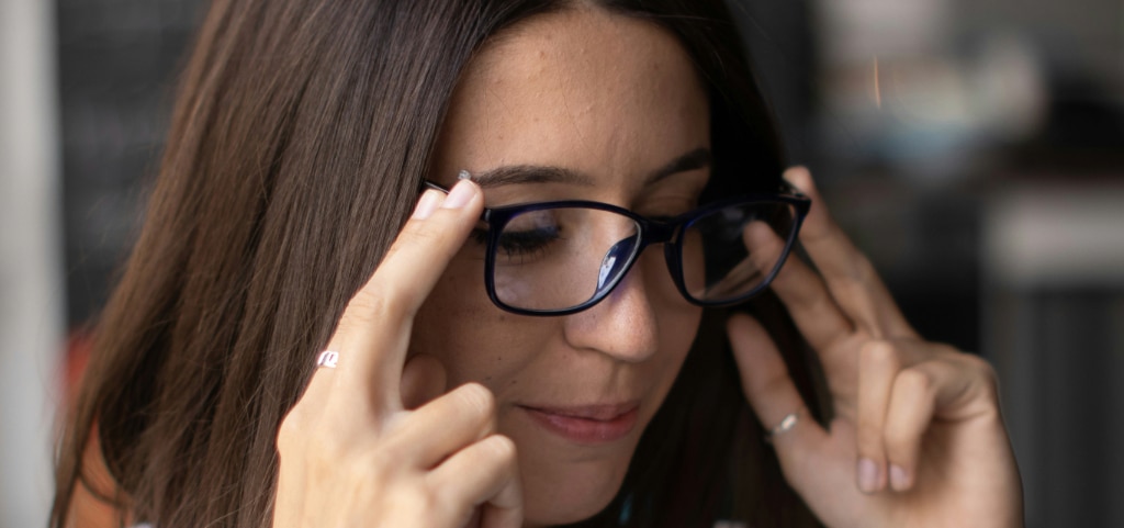 A close up of a young lady with long dark hair. She appears to be trying a pair of black acetate glasses on with both her index and forefingers grasping either side. 