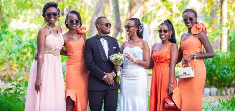 Bride and groom standing with bridesmaids outdoors in a bright summer setting, all wearing sunglasses; bridesmaids in peach and orange dresses, groom in a black tux and bride in a white strapless wedding gown holding a bouquet.