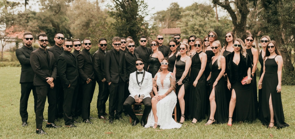 A large group photo of what appears to be wedding guests. The bride and groom are sitting at the centre. A group of men in black suits and sunglasses are standing to their right and a group of women in black dresses and sunglasses to their left. They are in a bright outdoor setting with trees in the background.