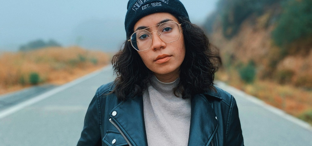 A close up of a young lady with dark curly hair and brown eyes. She is wearing a beanie, large aviator glasses, a leather jacket and grey jumper. She appears to be walking on a pathway surrounded by a mountainous landscape which is blurred slightly in the background.