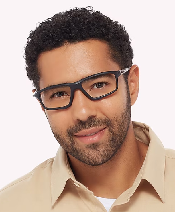 Close-up of a man with short dark curly hair wearing black rectangular Oakley wraparound glasses, smiling slightly against a plain background.
