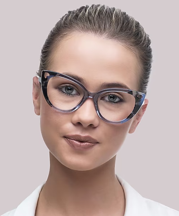 Close-up of a woman wearing blue Havana cat-eye glasses and a white top, with light hair pulled back and a neutral expression against a plain background.