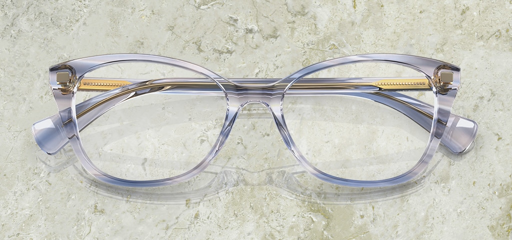 A pair of of acetate pillow-shaped Ralph by Ralph Lauren glasses. They have a transparent blue aesthetic with golden inner temples. The glasses are folded up on a marble background.