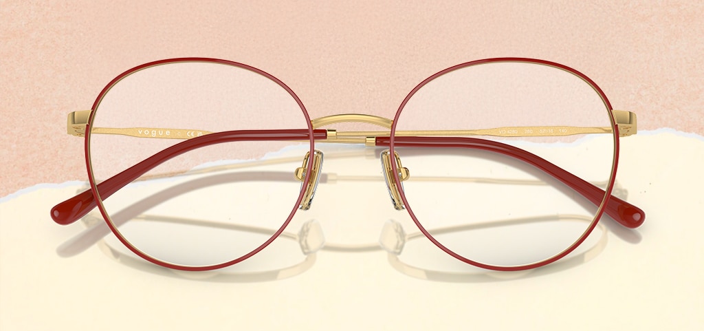 A pair of round, metal Vogue glasses propped open on a paper-torn peach and cream background. The glasses are a gold and red colour with the brand logo adorned along the temples.