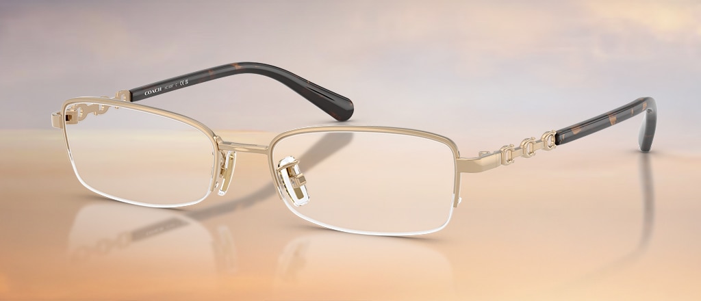 A pair of small rectangular semi-rimless glasses, made of gold metal with transparent nose pads on a gradient baby blue, white and peach background. The temples have a horseshoe-shaped interlocking design and black acetate tips. The glasses are folded up.
A pair of pillow-shaped Coach glasses at an angle on a gradient baby blue, white and peach background. The glasses are black with gold branded temples. The hinges have a unique floral design.