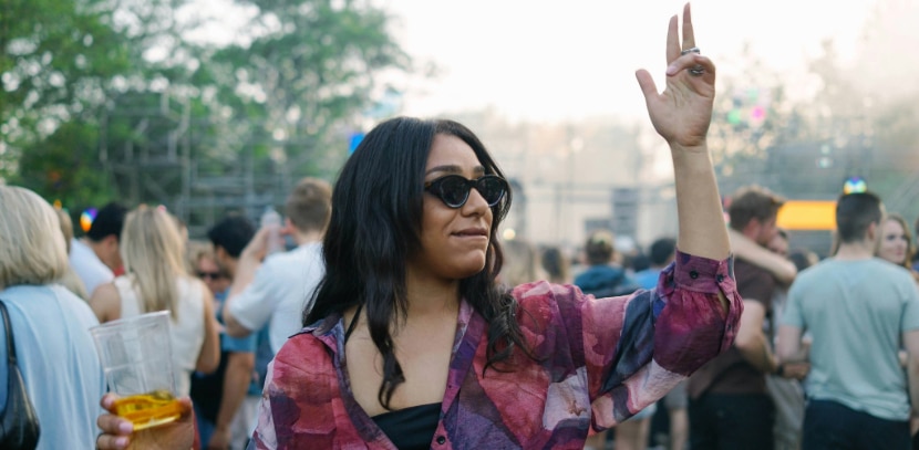 A woman with dark hair wearing black cat-eye sunglasses and a sheer patterned shirt over a black vest stands in a crowd at an outdoor summer festival, holding a drink in a plastic cup in one hand and raising the other as if dancing, with people and greenery blurred in the background.