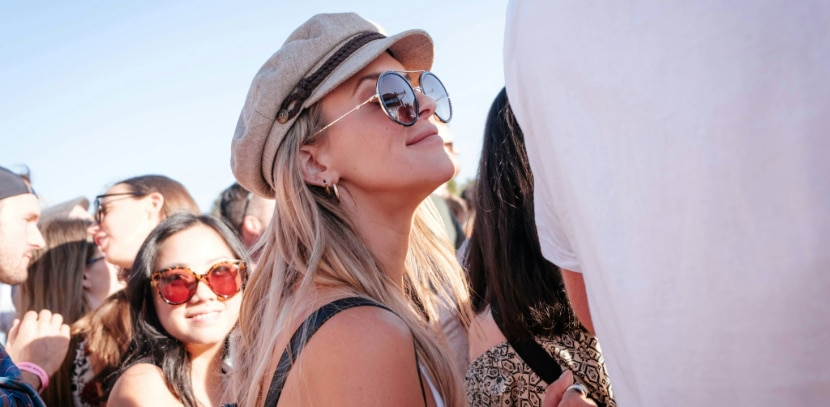 A woman with long blonde hair wearing a beige retro cap and large round sunglasses tilts her head upward, smiling in a crowd at an outdoor summer festival, with other people in sunglasses around her.
