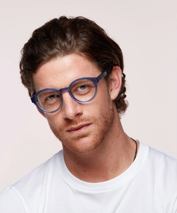 A man with medium-length wavy brown hair and light facial hair wears large round blue glasses with a white t-shirt, tilting his head slightly with a relaxed expression against a plain background.